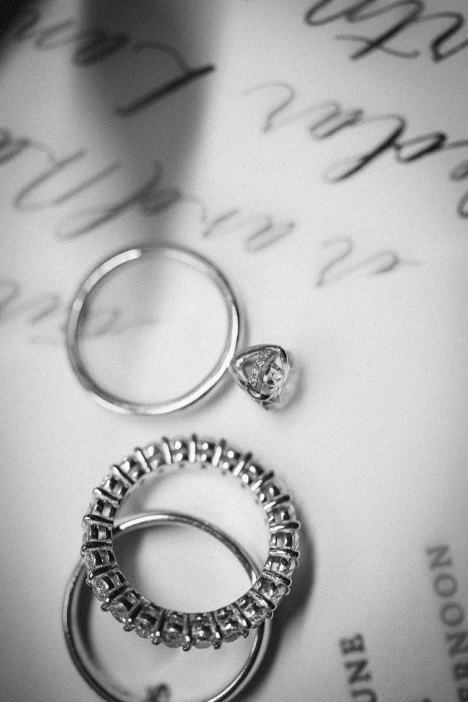 A black and white macro photograph of three wedding rings, including a diamond engagement ring and a pavé eternity band, resting on a wedding invitation with elegant cursive calligraphy; a perfect detail shot for a wedding photography side hustle. Photo by Susan Stripling.