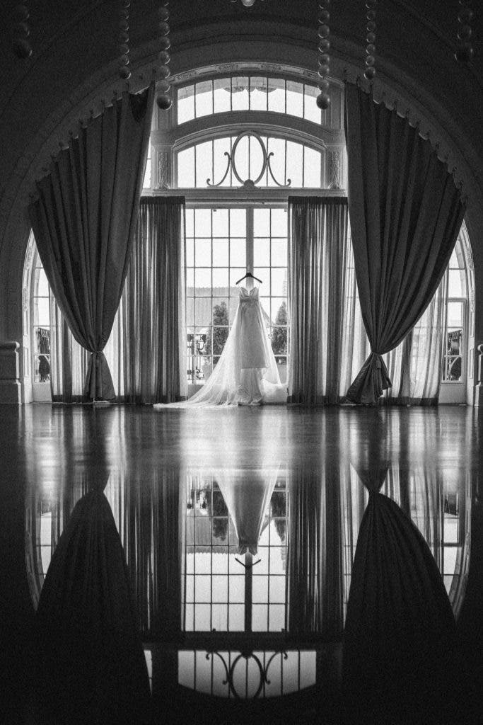 A dramatic black and white architectural shot of a bridal gown hanging centered in a massive arched window, with the entire scene perfectly reflected in the polished ballroom floor—an artistic composition for a wedding photography side hustle. Photo by Susan Stripling.