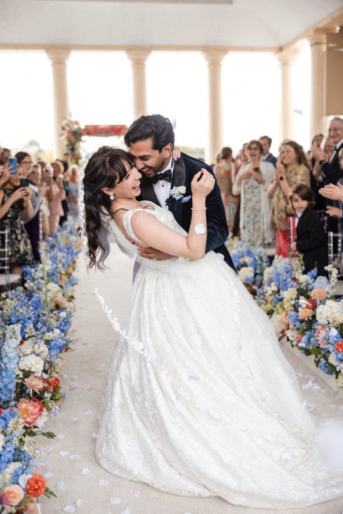 A vibrant color photograph of a groom dipping a laughing bride during their wedding recessional, surrounded by cheering guests and an aisle lined with lush blue and white flowers—a high-energy moment for a wedding photography side hustle. Photo by Susan Stripling.