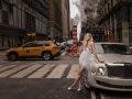 An editorial-style wedding portrait of a bride in a white fringe dress leaning against a luxury sedan on a busy New York City street with a yellow taxi in the background, demonstrating the urban flair of a wedding photography side hustle. Photo by Susan Stripling.