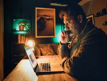 A man sits at a wooden table in a dimly lit, moody room, staring intently at a laptop screen with his chin resting on his hand. The scene is illuminated by the warm, orange glow of a desk lamp and a teal-colored backlight, creating a cinematic and focused atmosphere.