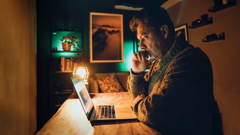 A man sits at a wooden table in a dimly lit, moody room, staring intently at a laptop screen with his chin resting on his hand. The scene is illuminated by the warm, orange glow of a desk lamp and a teal-colored backlight, creating a cinematic and focused atmosphere.