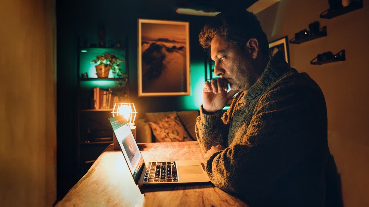 A man sits at a wooden table in a dimly lit, moody room, staring intently at a laptop screen with his chin resting on his hand. The scene is illuminated by the warm, orange glow of a desk lamp and a teal-colored backlight, creating a cinematic and focused atmosphere.