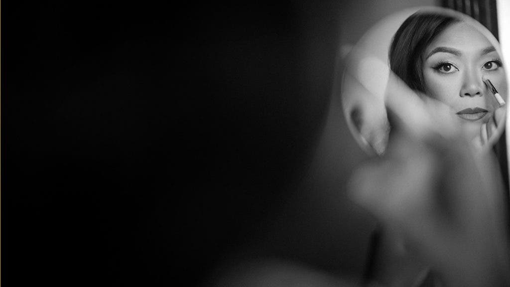 a bride gazes into the mirror as she applies undereye makeup, black and white photo