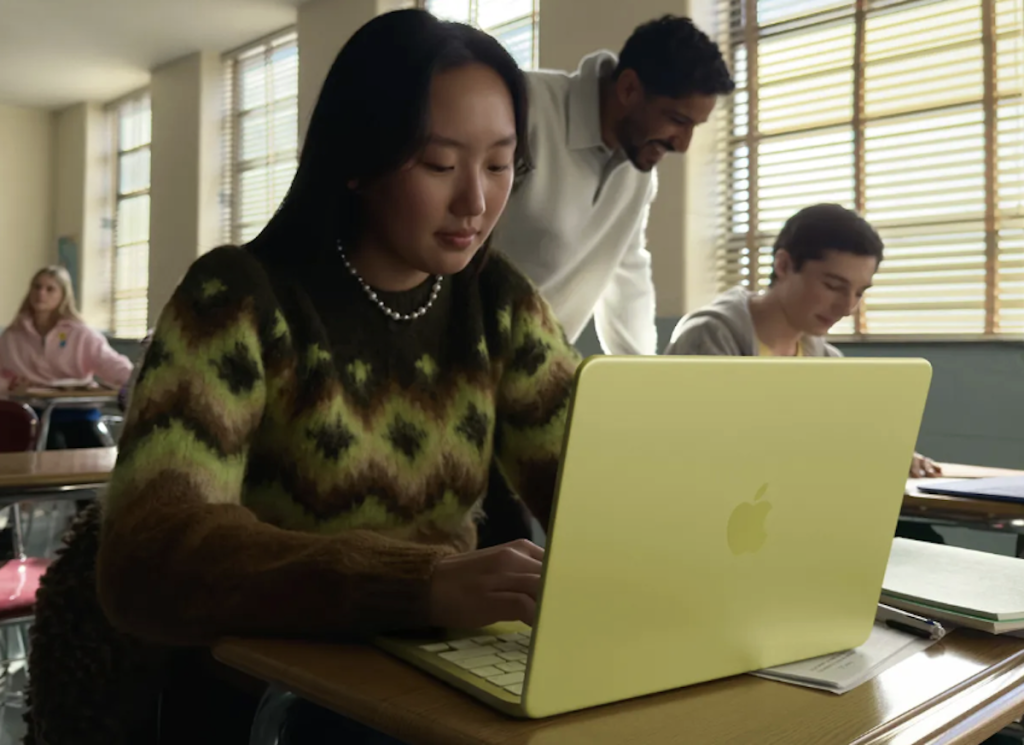A young woman with long dark hair, wearing a green and brown patterned sweater and a pearl necklace, works on a vibrant lime-yellow MacBook in a sunlit classroom. Behind her, a male teacher in a white sweater smiles while looking at another student's work.