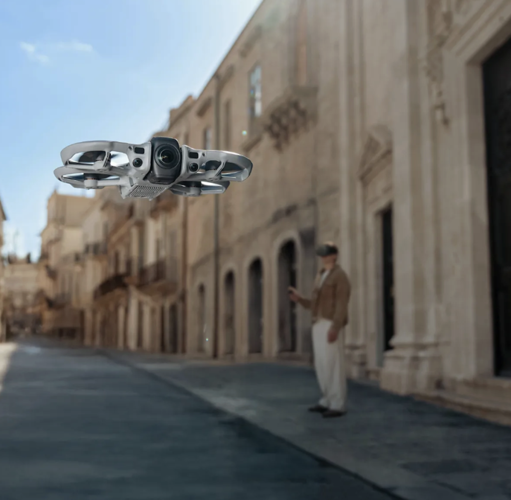 A small, white DJI Avata drone with protected rotors hovers mid-air in a narrow, historic European street with stone buildings. In the blurred background, a person stands on the sidewalk wearing a VR headset to pilot the drone.