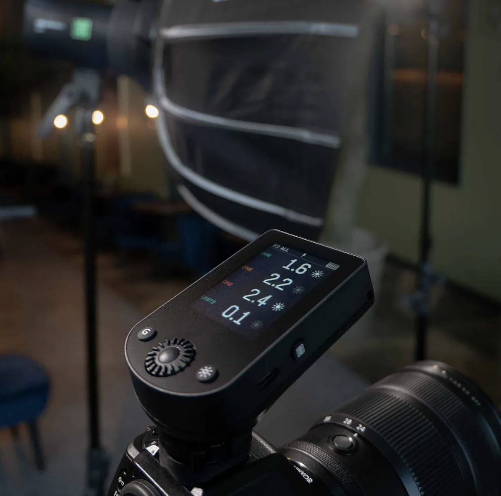 A close-up, low-angle shot of an Elinchrom Transmitter Pro X flash trigger mounted on the hot shoe of a Nikon camera. The touchscreen display is lit up, showing power levels for four different light groups (1.6, 2.2, 2.4, and 0.1) in a dark studio setting.