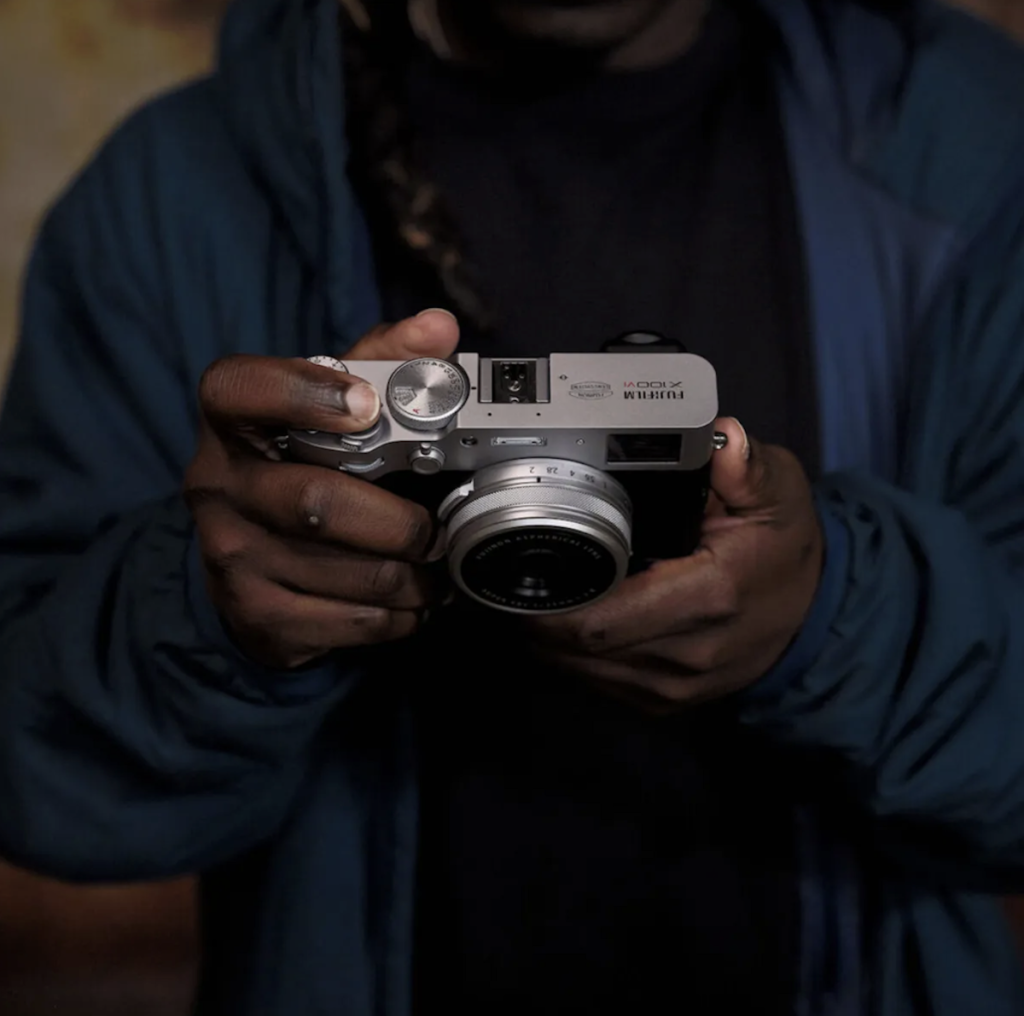A close-up of a person’s hands holding a silver and black Fujifilm X100VI digital camera at chest level. The focus is on the top of the camera, showing the manual shutter speed and exposure compensation dials.