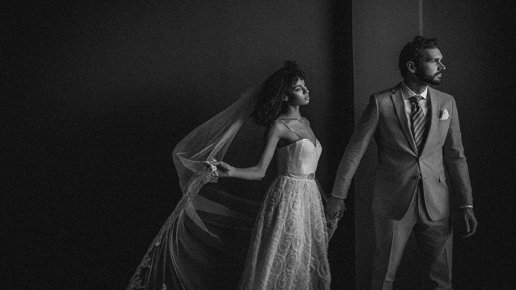 a bride and groom pose in a black and white photo, with the bride holding her veil 