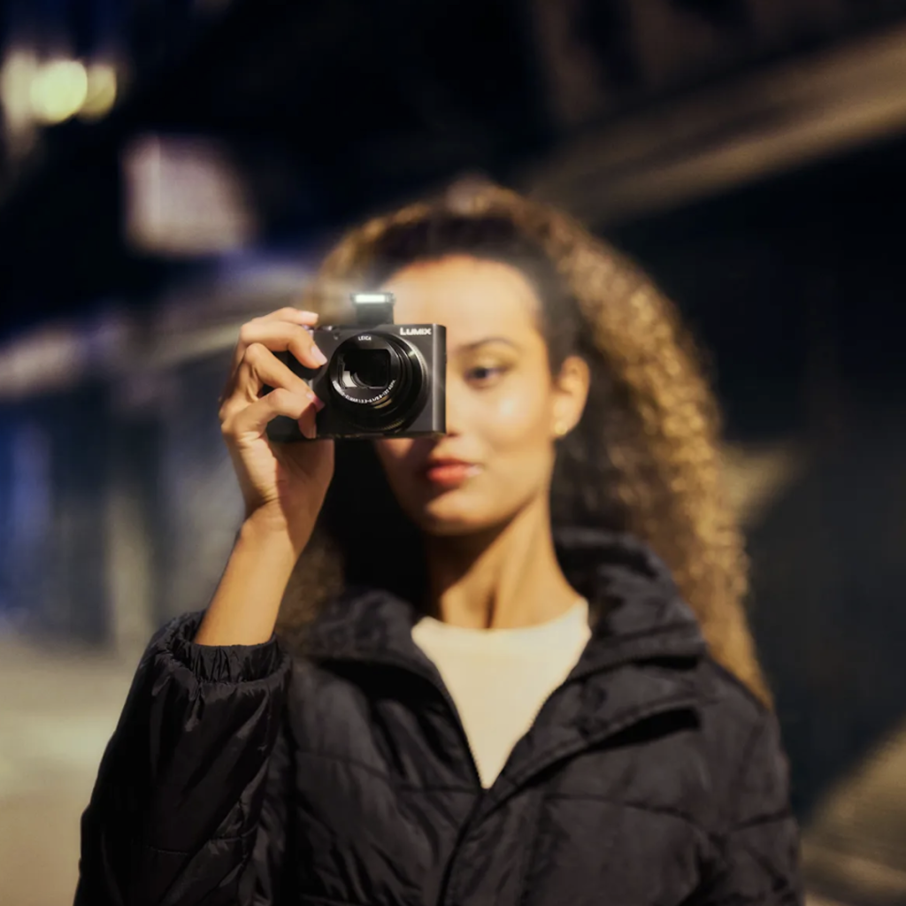 A woman with curly hair takes a photo at night, holding a black compact Panasonic LUMIX ZS300 camera to her eye. The camera’s built-in flash is firing, creating a bright burst of light against the dark, bokeh-filled city background.
