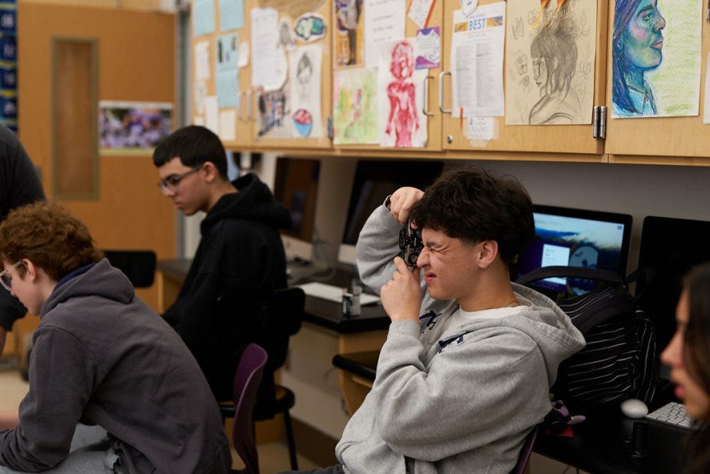 A high school student in a grey hoodie sits in a classroom, squinting as he looks through the viewfinder of a Pentax 17 camera, practicing the basics of analog photography.