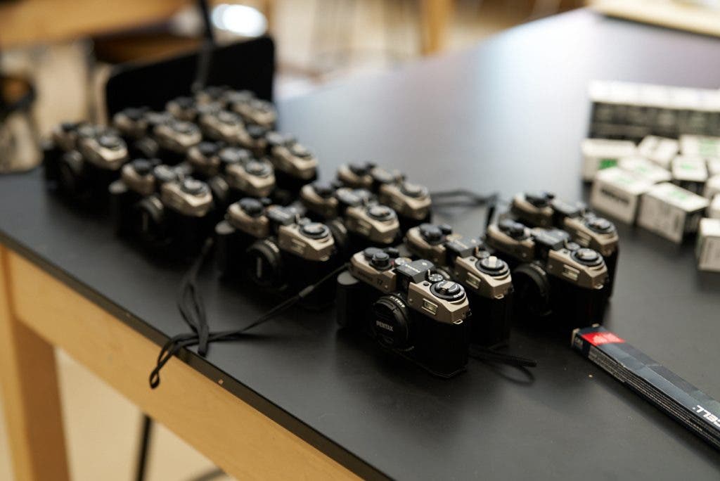 A fleet of several silver and black Pentax 17 cameras are lined up on a black table next to boxes of film, prepared for a workshop on analog photography.