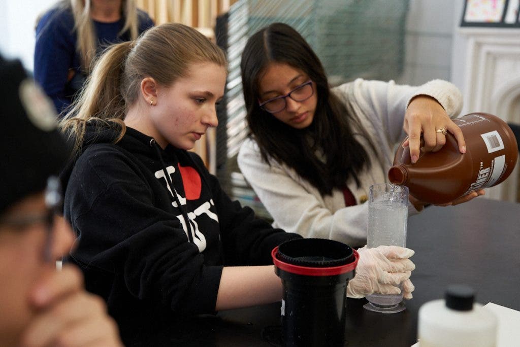 Two students focus intently on chemistry, with one pouring developer from a large brown jug into a graduated cylinder to process film for an analog photography project.