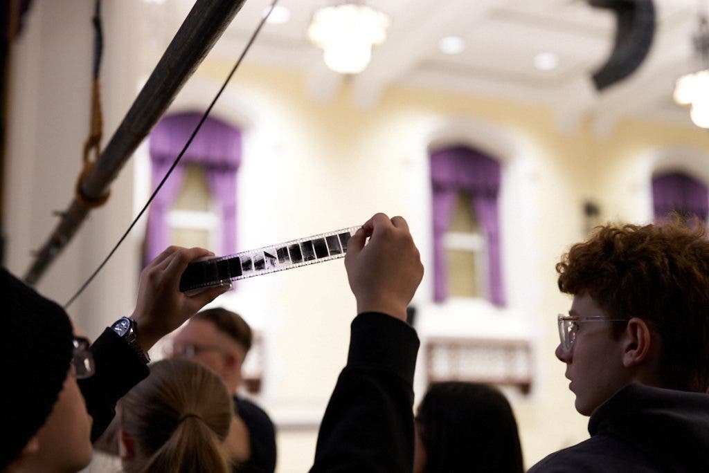 A student holds a strip of developed 35mm film negatives up toward the ceiling lights to inspect the captured frames, a rewarding moment in the analog photography process.