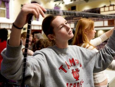 A student in a grey sweatshirt holds a long, translucent strip of processed film negatives across her field of vision, carefully reviewing her work during an analog photography workshop.