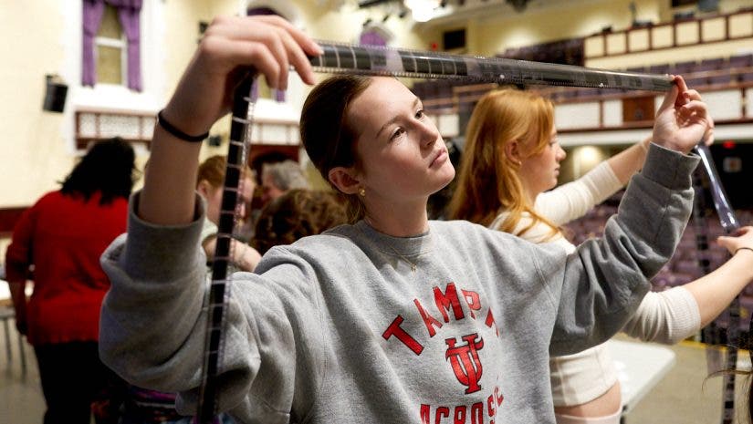 A student in a grey sweatshirt holds a long, translucent strip of processed film negatives across her field of vision, carefully reviewing her work during an analog photography workshop.