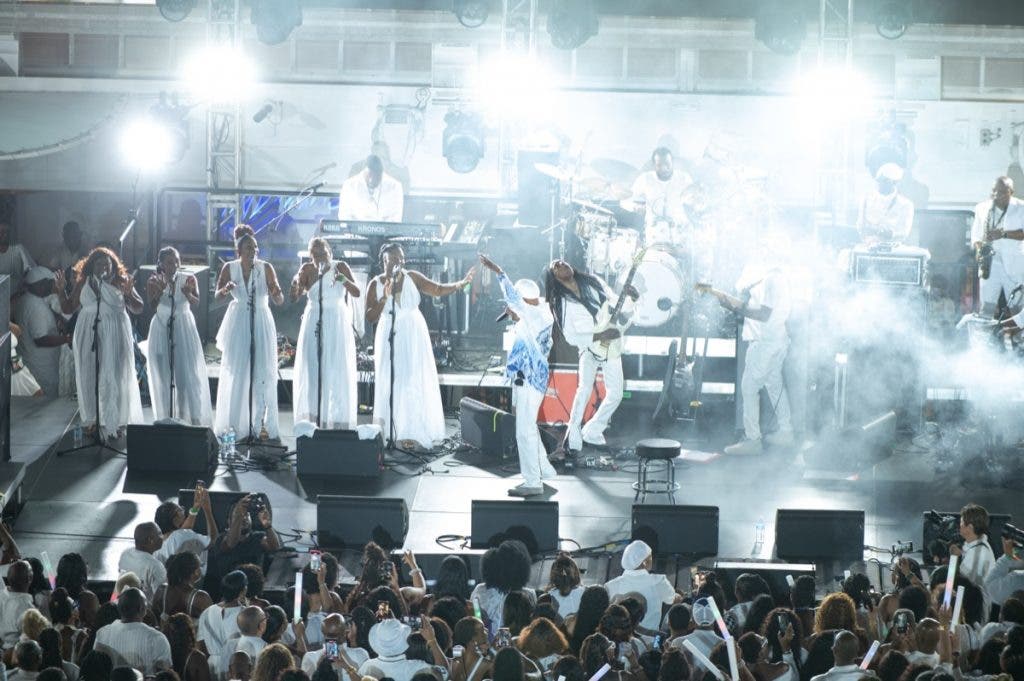 A live performance shot of a band and vocalists on stage, all dressed in white. The lighting is a mix of intense stage beams and atmospheric fill likely provided by Flashpoint speedlights, capturing the performers in a high-key, energetic glow while silhouetting the fans in the foreground.