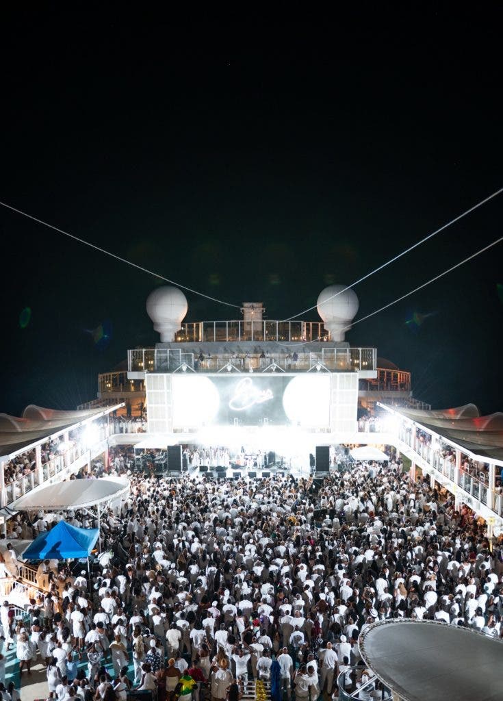 A high-angle, vertical wide shot looking down from an upper deck onto a massive "All-White" party on the pool deck of a cruise ship. Thousands of people in white clothing fill the deck space between the ship’s structures. Large glowing orbs and a massive LED screen displaying a logo illuminate the stage area at the far end.