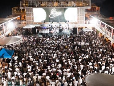 A wide, elevated landscape view of the same cruise ship deck party. The image captures the scale of the event, showing the crowd packed from the stage to the back of the deck. Bright white stage lights and overhead fixtures illuminate the scene, contrasting sharply with the dark ocean and sky surrounding the ship.