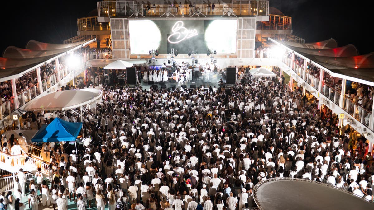 A wide, elevated landscape view of the same cruise ship deck party. The image captures the scale of the event, showing the crowd packed from the stage to the back of the deck. Bright white stage lights and overhead fixtures illuminate the scene, contrasting sharply with the dark ocean and sky surrounding the ship.