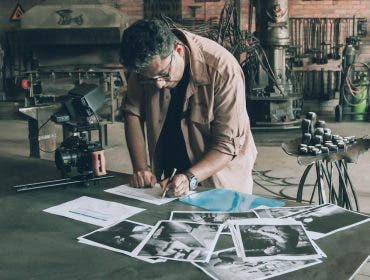 a man is looking down on a desk writing his shot list with photos scattered