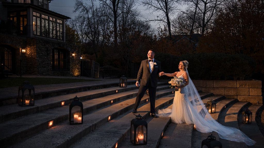 A bride and groom pose on wide stone steps at twilight, surrounded by glowing lanterns with a lit stone manor in the background; a romantic scene often shared by word of mouth, photographed by Susan Stripling.