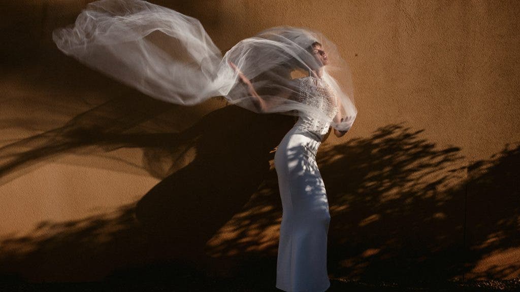 A dramatic portrait of a bride in a beaded gown with her long white veil billowing in the wind against a warm-toned wall and sharp shadows; a striking image known by word of mouth, photographed by Susan Stripling.