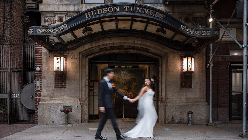 A cinematic motion-blur photo of a bride and groom walking past the historic "Hudson Tunnels" entrance in New York City; a classic urban wedding moment praised via word of mouth, photographed by Susan Stripling.