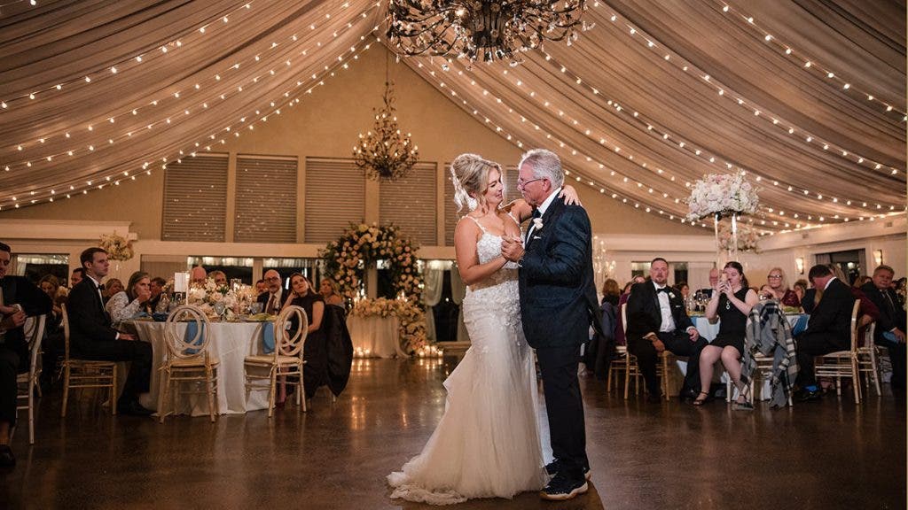 A bride shares an emotional dance with an older man in a grand reception hall featuring draped fabric ceilings and warm string lights; a heartfelt moment captured for word of mouth, photographed by Susan Stripling.