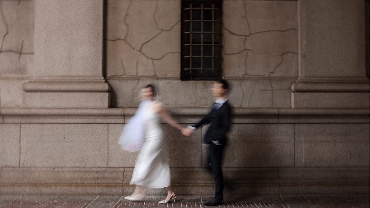 An artistic, motion-blurred shot of a bride and groom holding hands and walking quickly past a textured stone wall; a dynamic style that generates word of mouth, photographed by Susan Stripling.