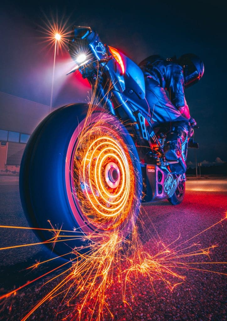 Dynamic long-exposure, low-angle shot from behind a motorcycle at night. Bright golden sparks spin aggressively off the rear tire, creating concentric fiery rings and scattering across the asphalt. The rider, clad in dark gear and a helmet, leans forward into the frame under a dramatic mix of blue, red, and neon street lighting. Created by Jordi Koalitic.