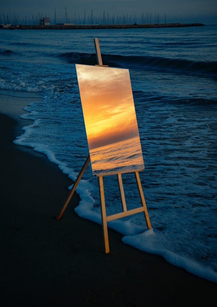 A wooden painter's easel stands on a sandy beach as a gentle ocean wave washes over its base. Instead of a canvas, a rectangular mirror rests on the easel, perfectly reflecting a vibrant, golden sunset over calm water. The surrounding beach and ocean are cast in deep, cool twilight blue, contrasting sharply with the warm reflection. Created by Jordi Koalitic.