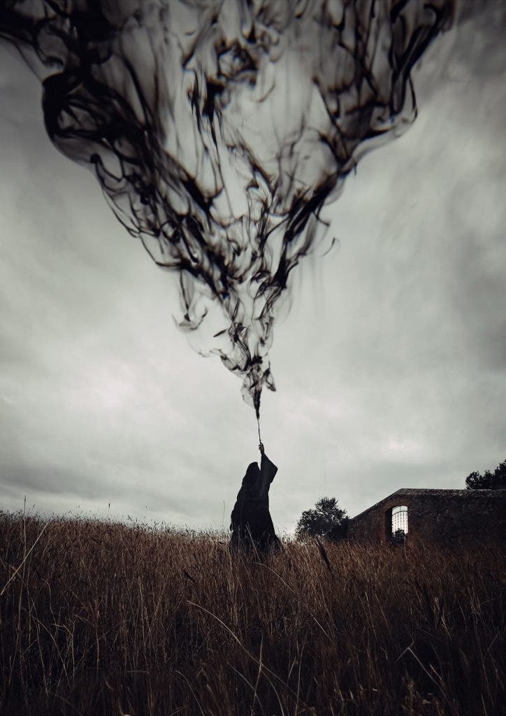 A surreal, dramatic shot of a dark, hooded figure standing in a field of tall, dry grass beneath an overcast sky. The figure holds up a wand or stick, from which an immense plume of black smoke erupts upward, swirling and expanding heavily into the sky like dark ink in water. An old stone building with a single window is visible in the background. Created by Jordi Koalitic.