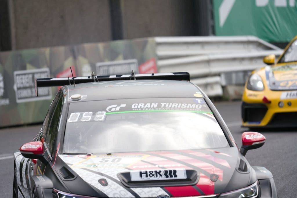 Sharp close-up tracking shot of the front windshield and roof of a moving black and red race car bearing a Gran Turismo sunstrip, with another yellow car blurred in the background.