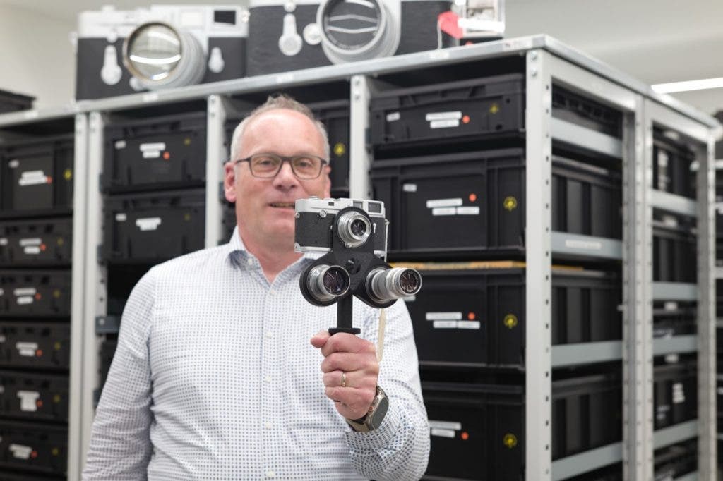 A man with glasses smiling and holding up a vintage silver Leica camera equipped with a three-lens turret attachment, standing in front of shelves filled with black storage cases.