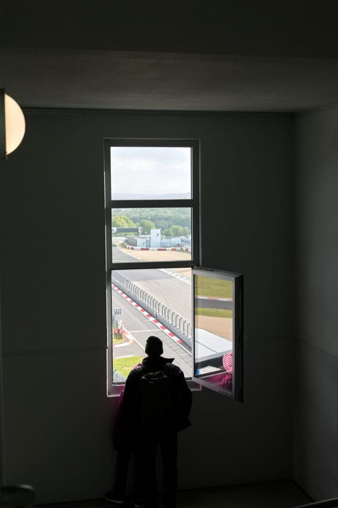 High-contrast silhouette of a person looking out of a tall, narrow open window inside a dark room, framing a view of a curving racetrack exterior.