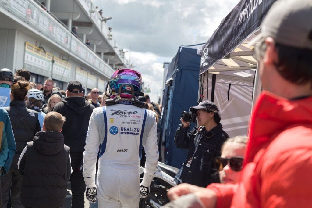 A crowded racetrack paddock under a bright sky, focusing on the back of a race car driver in a white racing suit with a colorful helmet walking past spectators and a photographer.
