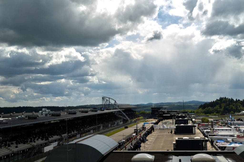 A wide-angle landscape view of a vast racetrack facility and grandstands under an expansive, dramatic sky filled with dark, heavy storm clouds and breaking light.
