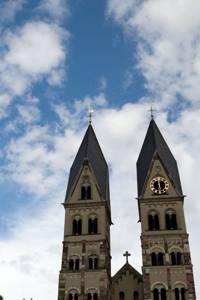 Low-angle shot looking up at the two tall twin stone spires of a historic European-style church with a golden clock face against a bright blue sky with white clouds.
