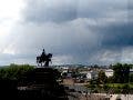 Dramatic wide shot of a dark silhouette monument of a person on a horse overlooking a riverfront city plaza under a massive, ominous storm cloud formation. Photographed with Leica SL3-P beta firmware by Daniel Norton