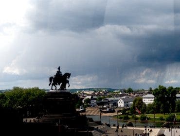 Dramatic wide shot of a dark silhouette monument of a person on a horse overlooking a riverfront city plaza under a massive, ominous storm cloud formation. Photographed with Leica SL3-P beta firmware by Daniel Norton