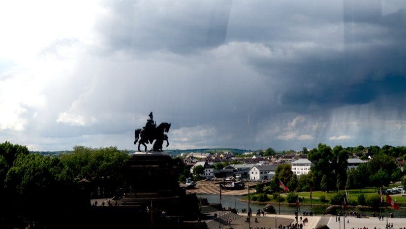 Dramatic wide shot of a dark silhouette monument of a person on a horse overlooking a riverfront city plaza under a massive, ominous storm cloud formation. Photographed with Leica SL3-P beta firmware by Daniel Norton