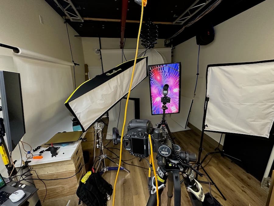 A photography studio setup featuring a camera mounted on a tripod pointing towards a vertical monitor, which displays a vibrant pink and purple geometric tunnel pattern. Two large rectangular softboxes are positioned on either side to light the scene.