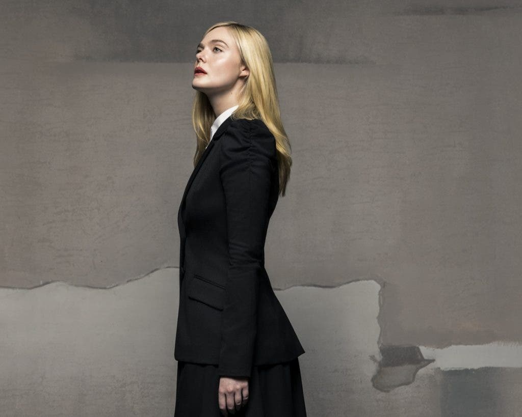 Side-profile portrait of actress Elle Fanning looking upward with a serene expression, wearing a tailored black suit jacket over a white collared shirt against a textured gray studio backdrop