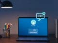 A laptop sits on a desk in a dimly lit room, illuminated by a black desk lamp. The screen displays a blue chatbot interface featuring a robot icon and a message reading, "Hello, Can I help You?", while a glowing, neon-style chat bubble graphic floats above the screen