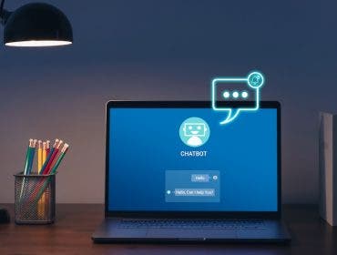 A laptop sits on a desk in a dimly lit room, illuminated by a black desk lamp. The screen displays a blue chatbot interface featuring a robot icon and a message reading, "Hello, Can I help You?", while a glowing, neon-style chat bubble graphic floats above the screen