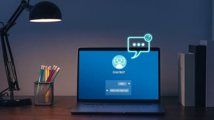 A laptop sits on a desk in a dimly lit room, illuminated by a black desk lamp. The screen displays a blue chatbot interface featuring a robot icon and a message reading, "Hello, Can I help You?", while a glowing, neon-style chat bubble graphic floats above the screen