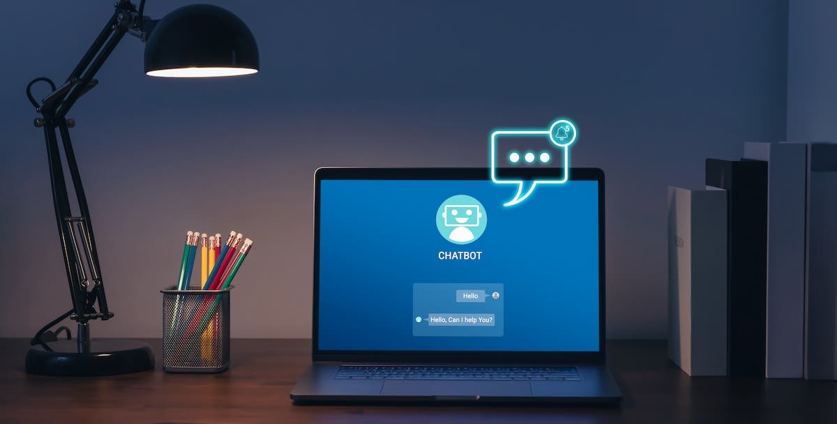 A laptop sits on a desk in a dimly lit room, illuminated by a black desk lamp. The screen displays a blue chatbot interface featuring a robot icon and a message reading, "Hello, Can I help You?", while a glowing, neon-style chat bubble graphic floats above the screen