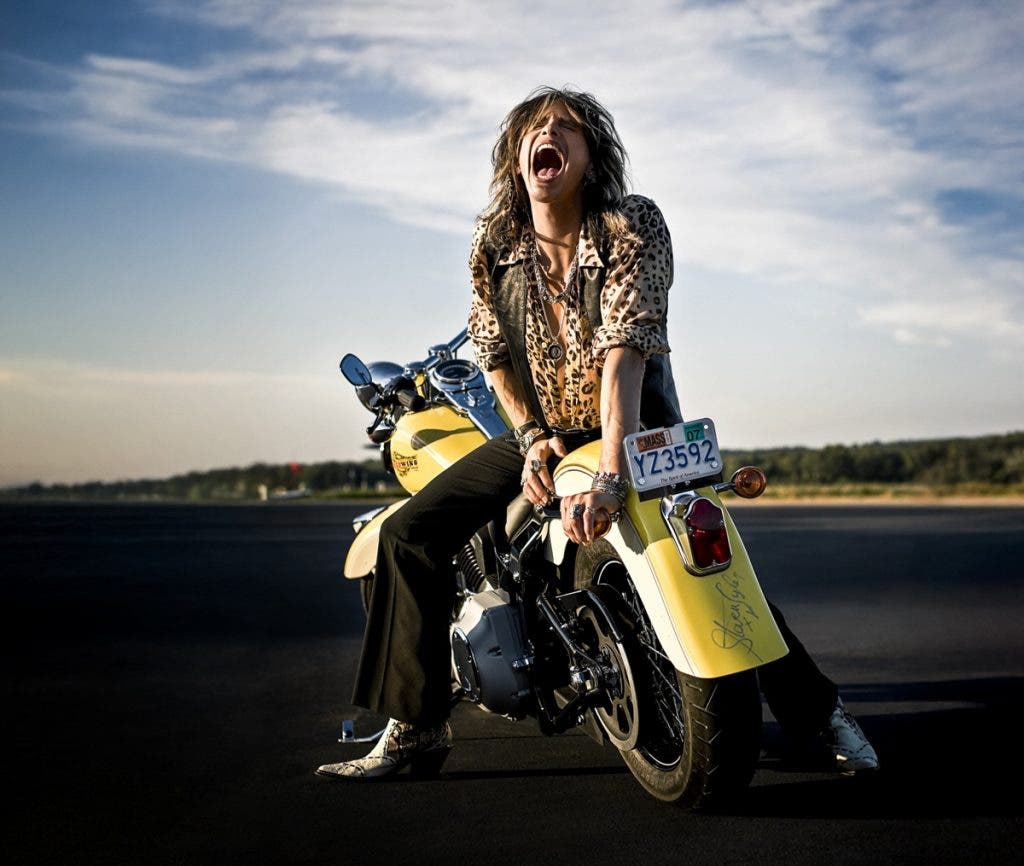 Steven Tyler sitting astride a bright yellow vintage motorcycle on an open asphalt strip, wearing a leopard-print shirt and leather vest while shouting dramatically with his head tilted back