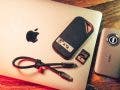 A silver Apple MacBook resting on a wooden desk. Placed on top of and beside the laptop are a SanDisk 2TB portable SSD labeled "MUDI 1", a 128GB SanDisk SD card, a short braided USB-C cable, and a gray llano-brand external hub or card reader